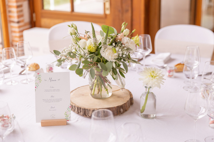 Centre de table de mariage : bouquet blanc et vert dans un bocal posé sur un rondin de bois, menu sur chevalet et soliflore