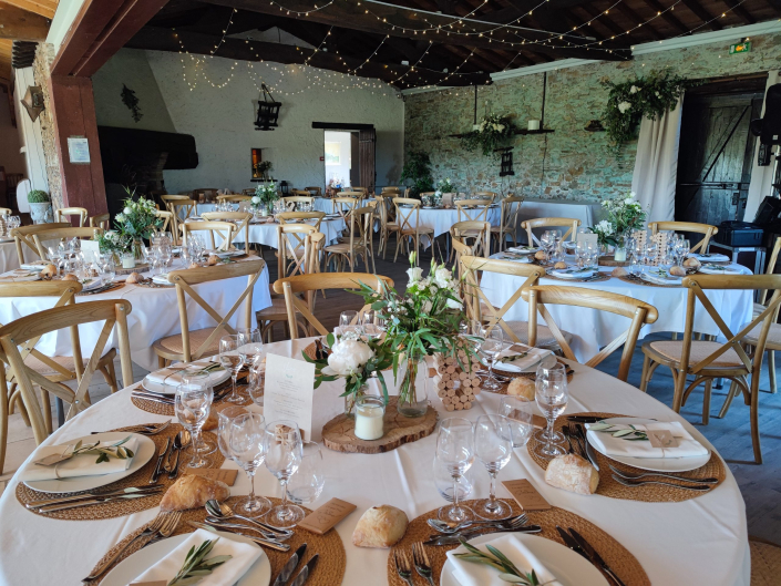 Salle de réception de mariage dans une grange : tables rondes, chaises en bois croisé, guirlandes lumineuses et centres de table végétaux