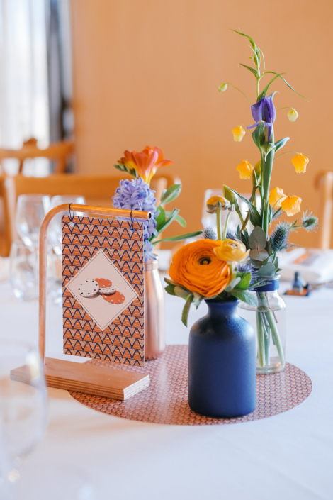 Table de mariage colorée : fleurs orange et violettes dans des vases bleus et numéro de table graphique sur support en bois