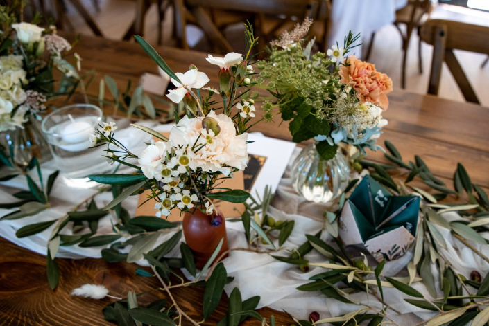 Centre de table champêtre aux branches d’olivier Centre de table champêtre : petits vases de fleurs blanches et pêche, branches d'olivier et photophore sur une table en bois