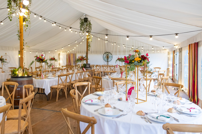 Mariage sous chapiteau : guirlandes guinguette, tables rondes, chaises en bois croisé et compositions de fleurs colorées sur colonnes dorées