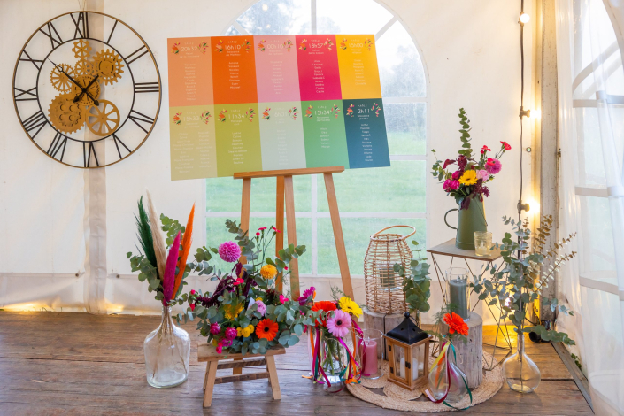 Plan de table multicolore sur chevalet en bois, horloge décorative, lanternes et bouquets de fleurs vives